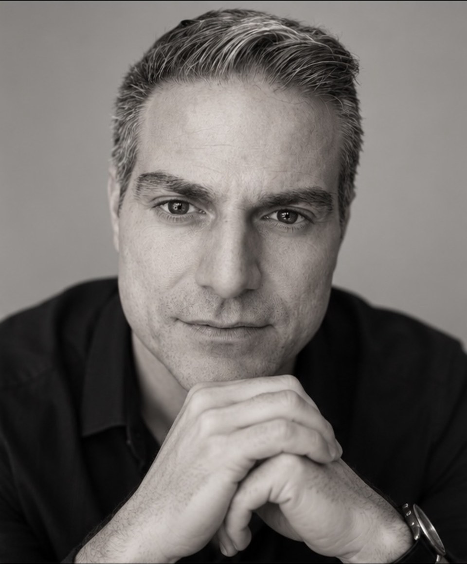 Andreas Aroditis in a black-and-white studio portrait, hands clasped beneath his chin, looking directly at the camera.