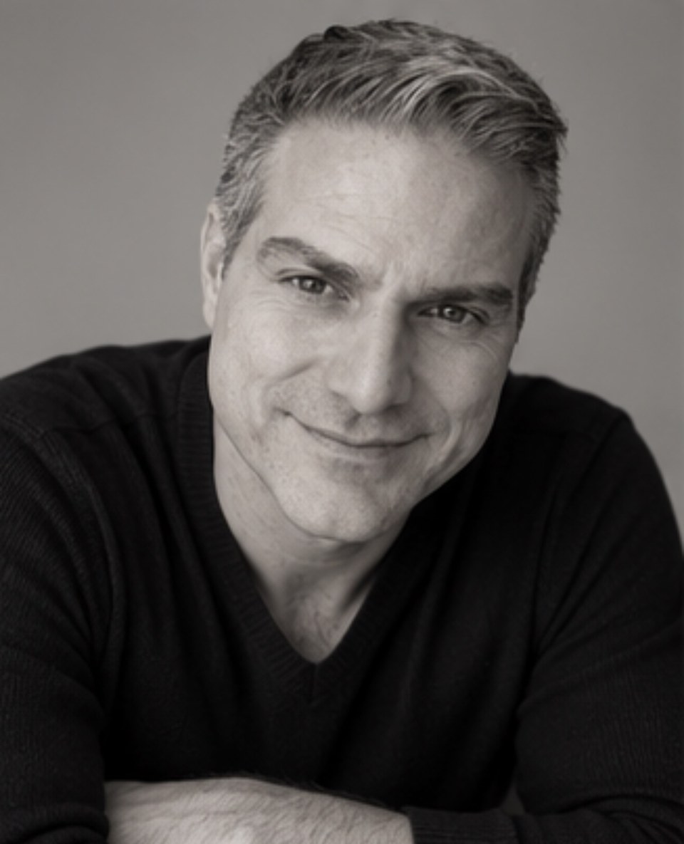 Andreas Aroditis in a black-and-white studio portrait, leaning forward with a half-smile.